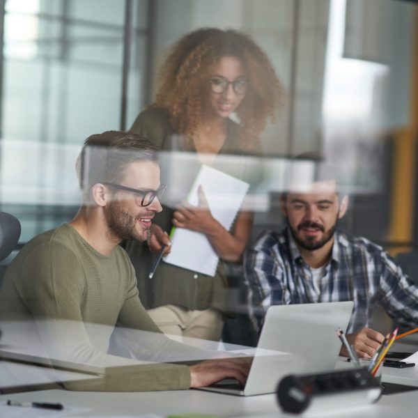 Helping coworkers. Three young happy colleagues using laptop and discussing work or sharing fresh ideas while working together in the modern office. Collaboration and teamwork. Brainstorming